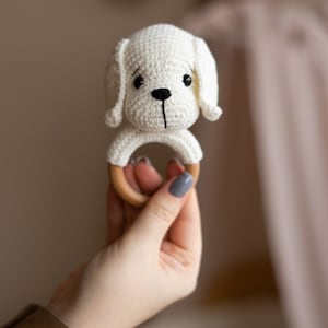 May include: A white crocheted dog rattle with a wooden ring. The rattle is held in a hand.