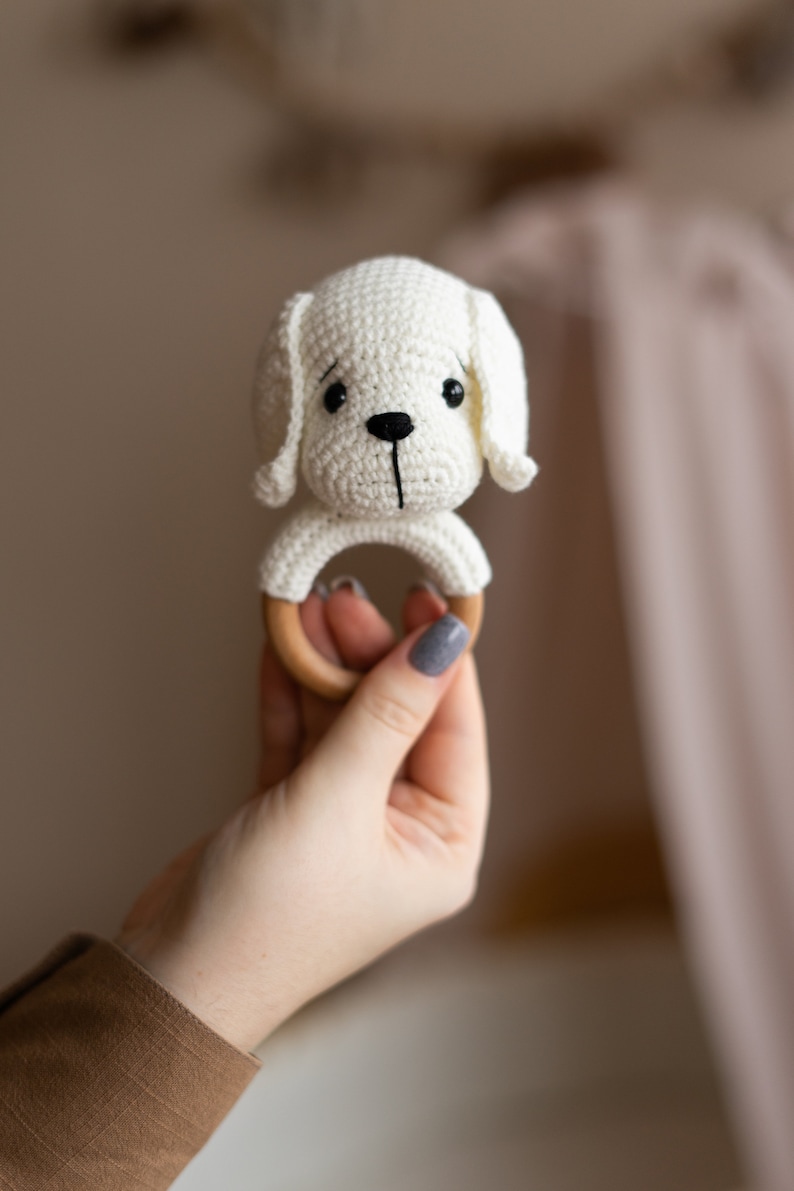 May include: A white crocheted dog rattle with a wooden ring. The rattle is held in a hand.