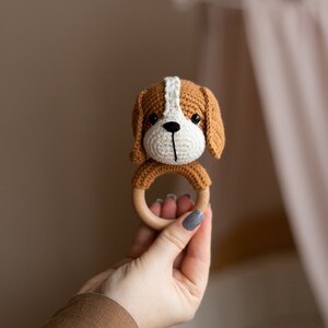May include: A crocheted brown and white dog rattle with a wooden ring. The rattle is held in a hand.