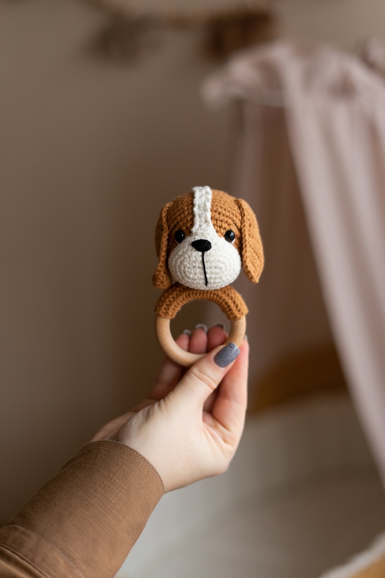 May include: A crocheted brown and white dog rattle with a wooden ring. The rattle is held in a hand.