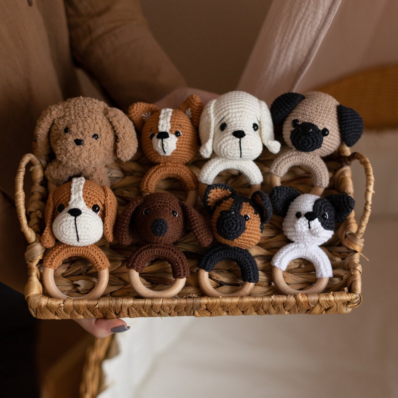 May include: A set of eight crocheted dog rattle toys in various colors and patterns. The toys are arranged in a wicker basket and feature different dog breeds, including a brown and white dog, a black and white dog, and a brown dog.
