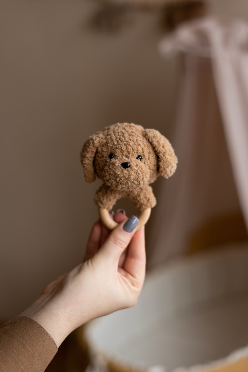 May include: A brown crocheted dog toy with a wooden ring. The toy is held in a hand with gray nail polish.
