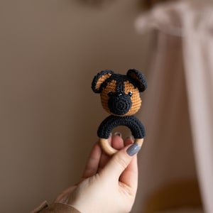 May include: A crocheted dog rattle with a wooden ring. The dog is brown and black with a black nose and eyes.