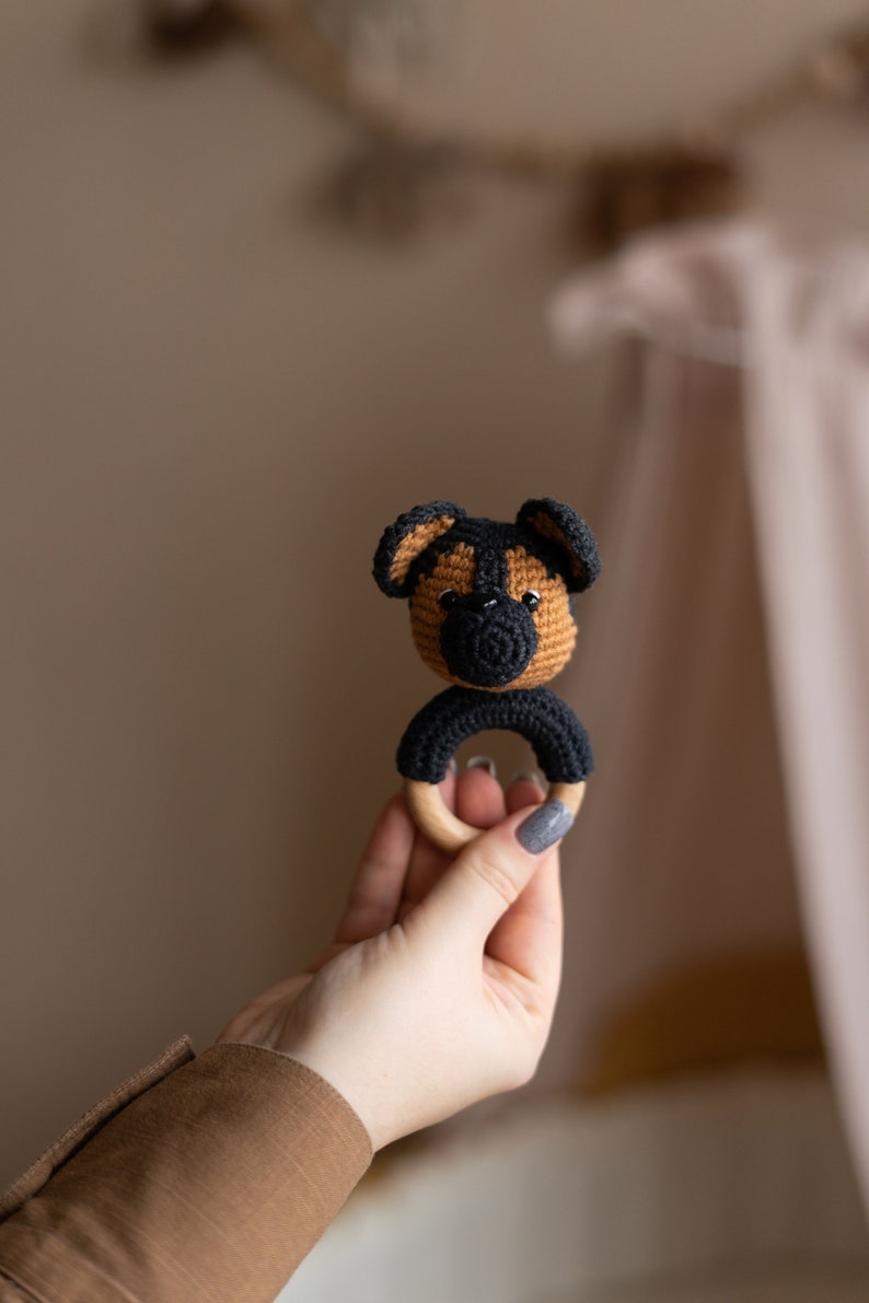 May include: A crocheted dog rattle with a wooden ring. The dog is brown and black with a black nose and eyes.