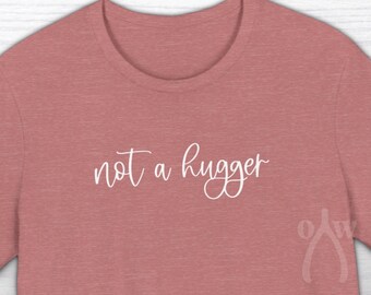 May include: A heather pink t-shirt with the text "not a hugger" in white cursive writing.