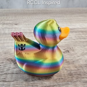 Op de afbeelding: Een regenboogkleurig eendenbeeldje met een oranje snavel en zwarte ogen. De eend heeft een goudkleurig gedeelte met een logo en een bundel roze items. De tekst "RCCL Inspired" staat bovenaan.