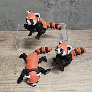 May include: Three red panda figurines. Each figurine has a red and white head, a black body, and a striped brown tail. One figurine is posed mid-jump, while the others are lying down. The figurines are displayed on a clear acrylic stand.