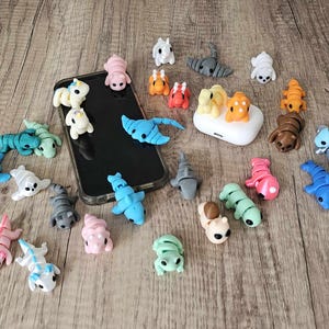 May include: A collection of colorful, small, animal-shaped figurines made of polymer clay. The figurines include a unicorn, a dragon, a scorpion, a turtle, a fish, a frog, a snail, a ladybug, a butterfly, a bee, a spider, a worm, a snake, a bird, and a cat. The figurines are arranged on a wooden surface.