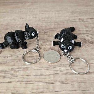 May include: Two black cat keychains with silver key rings. One cat is curled up and the other is lying on its belly.