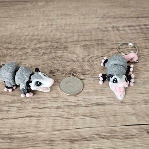 May include: Two gray and white possum keychain charms with pink accents. The possums are in different poses, one is curled up and the other is standing on its hind legs.