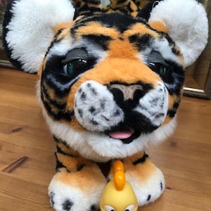 Pode incluir: Um brinquedo de pelúcia de tigre com pelo laranja, preto e branco, orelhas grandes e uma expressão amigável. Na frente do tigre, há um pequeno brinquedo de pintinho amarelo com bico e patas laranja. Os brinquedos estão em uma superfície de madeira.
