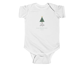 Puede incluir: Un body blanco de manga corta para bebé con un gráfico de un árbol de Navidad verde con una estrella dorada. Debajo del árbol, el nombre Léa está impreso en cursiva, seguido del texto MON PREMIER NOEL y el año 2026.