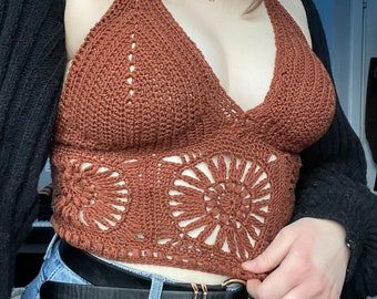May include: A brown crochet halter top with a floral design. The top is paired with blue jeans and a black belt with a gold buckle. A black cardigan is worn over the top.