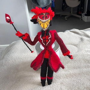 May include: A handmade crochet doll featuring a character with a red coat, black pants, and a yellow-toothed grin. The doll has a red hairstyle, black horns, and holds a red staff. The doll is approximately 10 inches tall.
