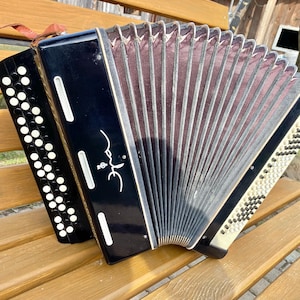 May include: A black and white accordion with a brown bellows and a silver trim. The accordion has a black leather strap and the words "Этюд" written on the side.