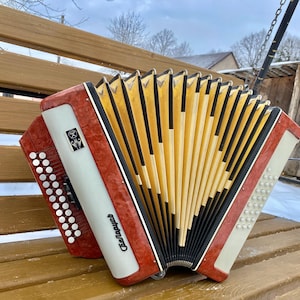 Pode incluir: Um acordeão vermelho e branco com um fole amarelo e preto. O instrumento tem um design decorativo na frente e está apoiado em um banco de madeira. O acordeão é um instrumento musical.