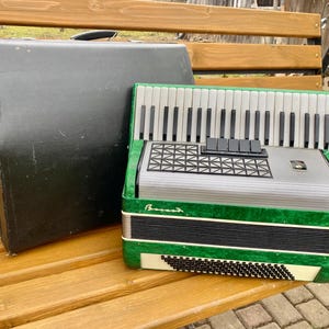 May include: A green and silver accordion with a black case. The accordion has a white keyboard and a black bellows. The case is closed and has a black handle.