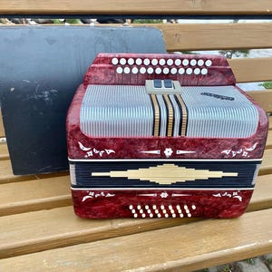 Pode incluir: Um acordeão vermelho e prateado com detalhes florais decorativos e uma caixa preta. O acordeão tem várias fileiras de botões brancos e uma grelha prateada. O instrumento está sobre um banco de madeira.