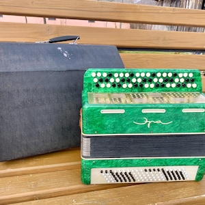 May include: A green accordion with black and white keys and buttons, and the word "Эра" in cursive script. A black case with a handle sits next to the accordion. The accordion is on a wooden bench.
