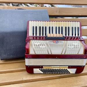 May include: A burgundy accordion with ivory and black keys, resting on a wooden bench next to a gray case. The accordion features a decorative grill and a logo. The instrument is designed for musical performance.