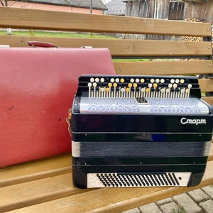 May include: A black and silver accordion with white and gold buttons, resting on a wooden bench next to a red carrying case. The accordion has the Cyrillic text "Старт" on its front.