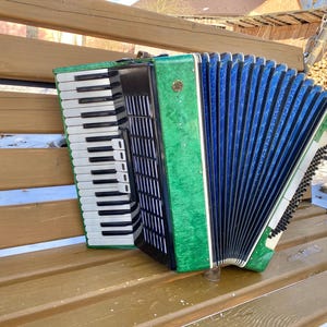 May include: A green and blue accordion with white and black keys. The instrument is resting on a wooden bench. The bellows are blue, and the keys are white and black. The accordion has a vintage look.