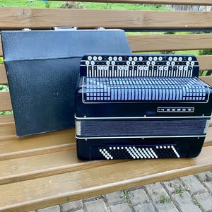 May include: A black accordion with white accents and a matching black case rests on a wooden bench. The accordion features a keyboard, buttons, and a bellows design. The case is rectangular and has a handle.