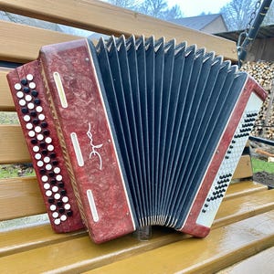 May include: A vintage red and black accordion with white and black buttons. The accordion is resting on a wooden bench. The bellows are black, and the instrument has a decorative design on the front.