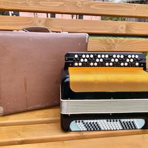 May include: A black and silver accordion with a yellow bellows and white and black keys. The accordion is sitting on a wooden bench next to a brown suitcase.