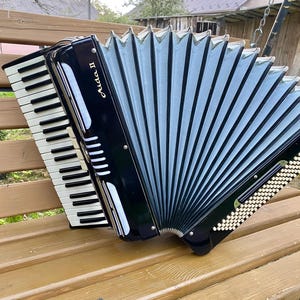 May include: A black and silver accordion with white and black keys, resting on a wooden bench. The accordion has the text "Aida II" on the front. The bellows are a light blue color.