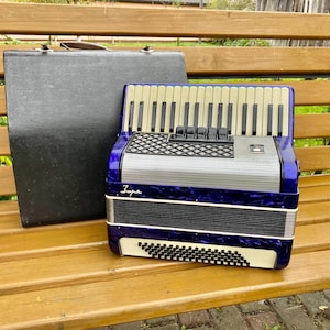 May include: A vintage blue and silver accordion with a black and white keyboard, resting on a wooden bench. A black carrying case is beside the accordion. The accordion has a decorative design and the word "Zupa" is written on it.