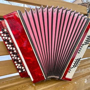 May include: A red and white accordion with a pink bellows, resting on a wooden bench. The instrument has a glossy finish and features multiple buttons and keys. The accordion is a musical instrument.