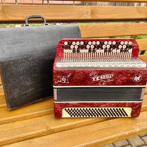 May include: A red accordion with black keys and a white button panel. The accordion has a black and white bellows and a black carrying case. The accordion has the word "TEMБР" written on the front.