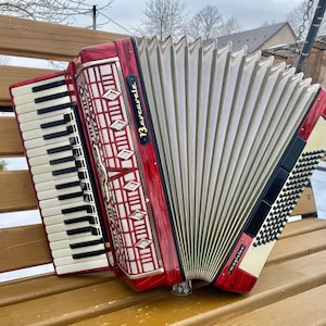May include: A red and silver accordion with a black and white keyboard. The accordion has the word "Barcarole" printed on it. The bellows are white and the instrument is resting on a wooden bench.