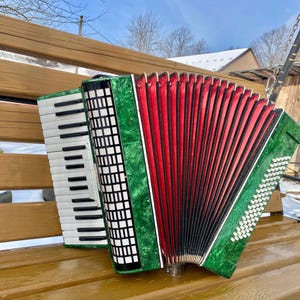 May include: A green, red, and black accordion with white keys and buttons rests on a wooden bench. The bellows are red, and the instrument has a decorative green trim. The accordion is positioned outdoors.
