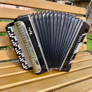May include: A black accordion with silver accents and white buttons rests on a wooden bench. The bellows are black and pleated, and the instrument has a rectangular shape. The name "Рубин" is visible on the front.