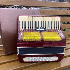 May include: A vintage burgundy accordion with ivory and black keys, yellow speaker covers, and a black and white grill. A brown carrying case is next to the accordion. The instrument is on a wooden bench.