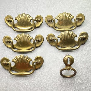 May include: Five antique brass drawer pulls and a keyhole escutcheon. The drawer pulls have a shell design with curved handles and ornate detailing. The keyhole escutcheon has a matching design. The hardware is likely for a dresser or cabinet.