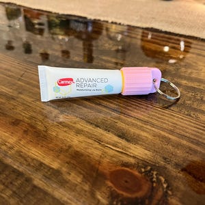 Könnte beinhalten: Eine rosa und weiße Tube Carmex Advanced Repair Feuchtigkeitsspendende Lippenpflege mit einem silbernen Schlüsselring am Deckel.