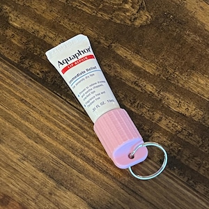以下が含まれることがあります： ピンクのキーホルダーに、小さなチューブのAquaphor Lip Repairバームが取り付けられています。チューブは白地に青色の文字で、「Aquaphor Lip Repair」と「Immediate Relief」と書かれています。