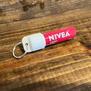 May include: A pink and white NIVEA lip care tube with a silver key ring attached. The tube says "NIVEA CHERRY LIP CARE" on it.