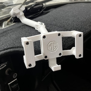 May include: A white 3D-printed phone mount for a car's dashboard. The mount features a rectangular frame with the letters "MG" in the center. The mount is attached to a flexible arm and is secured with black screws.