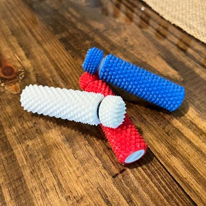 May include: Three cylindrical sensory tools with a textured, spiky surface. One tool is bright blue, one is solid white, and one is vibrant red. Each tool features a small, rounded end cap and a flat base, designed with a tactile, bumpy exterior.