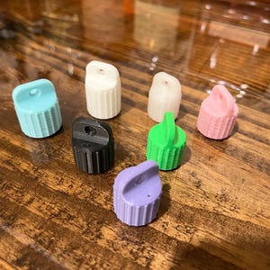 May include: Assortment of seven small plastic bottle caps in various colors, including light blue, white, pink, black, green, and purple. The caps have a small hole on top and a ribbed design.