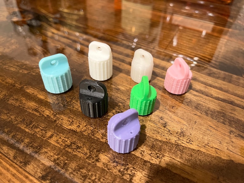 May include: Assortment of seven small plastic bottle caps in various colors, including light blue, white, pink, black, green, and purple. The caps have a small hole on top and a ribbed design.