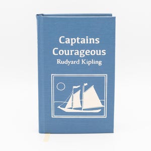 May include: A blue hardcover book titled "Captains Courageous" by Rudyard Kipling. The cover features a white illustration of a sailboat with a sun and a rectangular border. A small yellow bookmark is visible at the bottom.