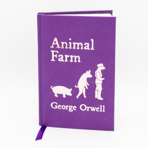 Puede incluir: Un libro de tapa dura morado con el título "Animal Farm" en letras blancas. La portada también muestra ilustraciones de un cerdo, un perro y una figura con sombrero. El nombre del autor, George Orwell, está impreso debajo.