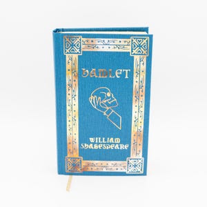 Op de afbeelding: Een teal hardcover boek getiteld "Hamlet" van William Shakespeare. De omslag heeft gouden decoratieve randen en letters, met een illustratie van een schedel in een hand. Een crèmekleurig bladwijzer is zichtbaar.