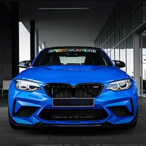 Op de afbeelding: Een felblauwe sportwagen met een zwarte grille, hoekige koplampen en een voorruitsticker met de tekst SPEEDHUNTERS in een holografische afwerking. Het voertuig heeft een lage voorbumper met zwarte luchtinlaatopeningen.