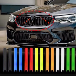Puede incluir: Una rejilla frontal de coche negra con un óvalo de puntos rojos, acompañada de un conjunto de catorce clips de inserción verticales de varios colores, incluyendo negro, azul, rojo, amarillo, naranja, blanco y verde, dispuestos en fila.